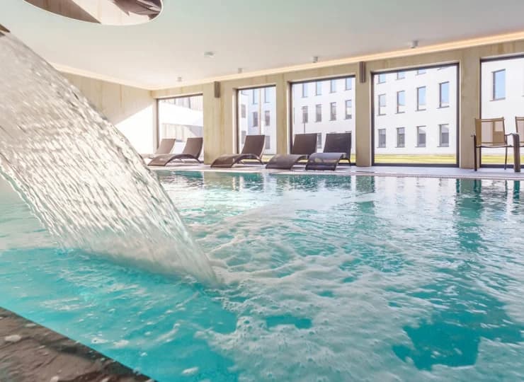 W basenie jest kaskada do hydromasażu, obok stoją leżaki
